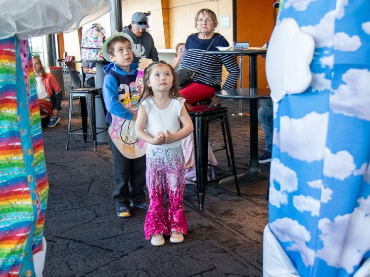 Image of Two young children stand indoors, looking up in awe at two people dressed in colourful clown costumes. Adults sit and watch in the background near a bar area with a toilets sign overhead.