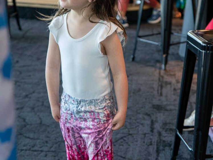 Image of A young girl with long brown hair, wearing a white top and sparkly pink mermaid-style trousers, stands indoors on a dark carpet, looking up. Other children and adults are blurred in the background.