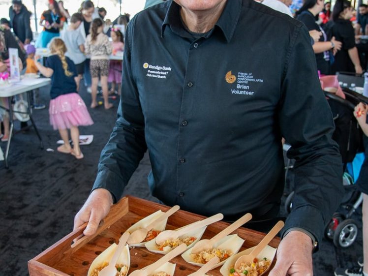 Image of A smiling man in glasses and a black shirt holds a wooden tray with several wooden spoons filled with food, standing in a busy indoor event space with people in the background.