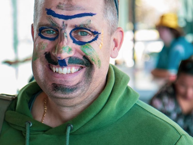 Image of A man wearing a green hoodie and sunglasses on his head smiles at the camera with colourful face paint, including blue, green, and black lines and shapes, in a brightly lit indoor setting.