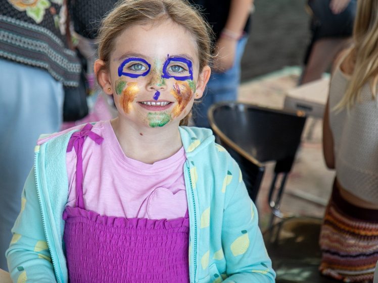 Image of A young girl with colourful face paint smiles at the camera. She is wearing a purple dress and a light blue jacket with yellow spots. There are people and chairs in the blurred background.