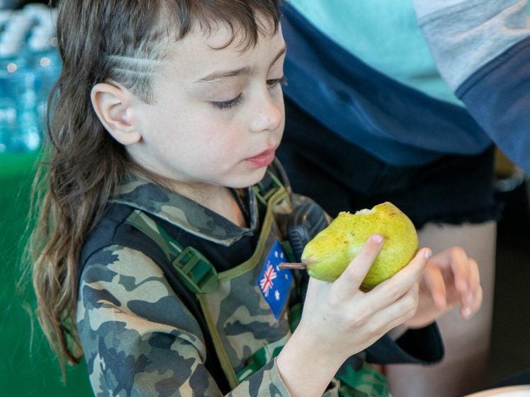 Image of A young child in camouflage clothing examines a large green pear, preparing to eat it whilst sitting at a table with other food items.