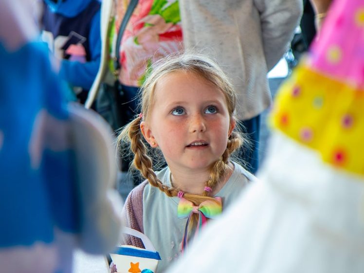 Image of A young girl with plaits holds a decorated cup and looks up with curiosity, surrounded by adults and children at what appears to be a festive event. Brightly coloured costumes are visible in the foreground.