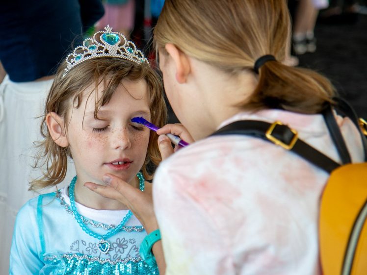 Image of A young child in a blue princess dress and tiara has her eyes closed whilst another person carefully applies blue face paint to her eyelid.