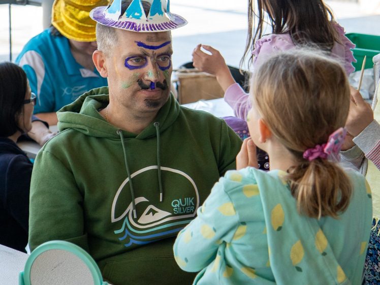 Image of A man wearing a handmade paper crown and face paint sits at a table, looking at a child in a yellow spotty hoodie. Other children and adults are engaged in arts and crafts activities around them.
