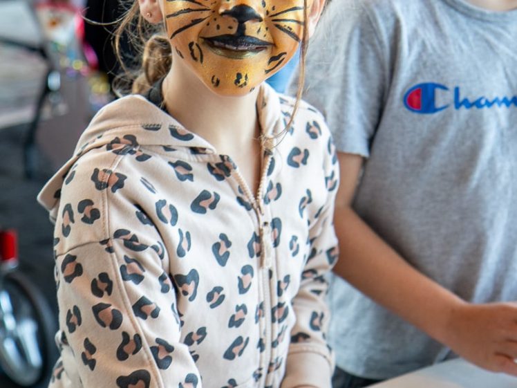 Image of A young child with their face painted like a tiger, wearing a leopard-print hoodie, stands at a table with art supplies, taking part in a craft activity. Another child in a grey shirt stands beside them.