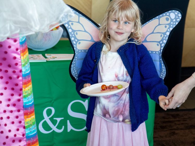 Image of A young girl wearing butterfly wings, a blue jumper, and a pink skirt holds a plate with snacks and smiles while holding an adult’s hand at an indoor event. A green tablecloth is visible in the background.