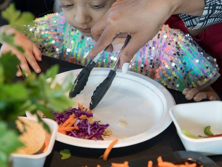 Image of A child in a colourful costume and mask watches as an adult uses tongs to place shredded vegetables onto a white plate at a table set with various fresh ingredients.