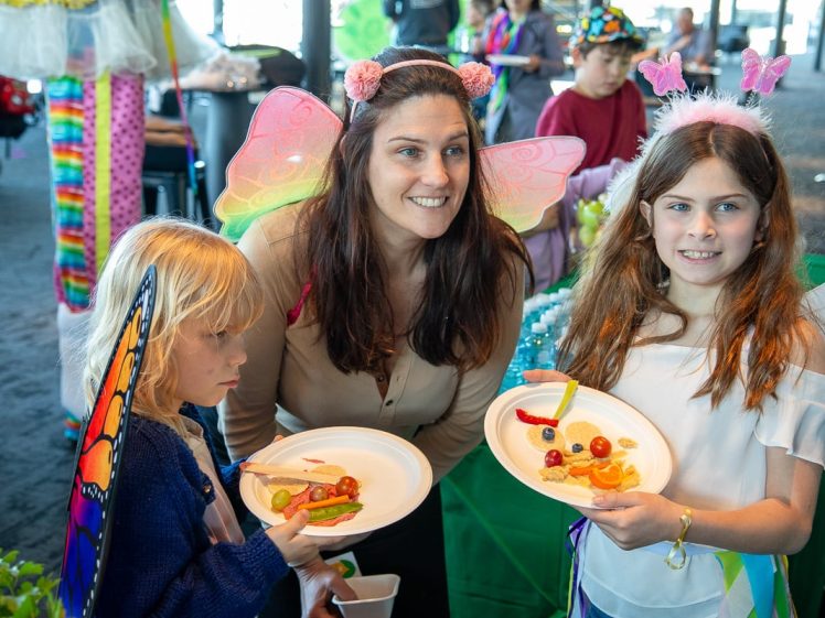 Image of Three people wearing fairy wings and headbands smile for a photo at a colourful indoor event. Two children hold plates of snacks shaped like insects and flowers. Other attendees and decorations are visible in the background.