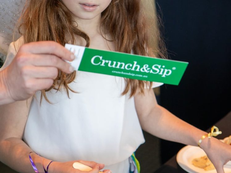 Image of A young girl spreads a topping on a biscuit at a table. In the foreground, a hand holds a green sign that reads “Crunch&Sip.” The girl is focused on her task, with more biscuits and dip on a plate nearby.
