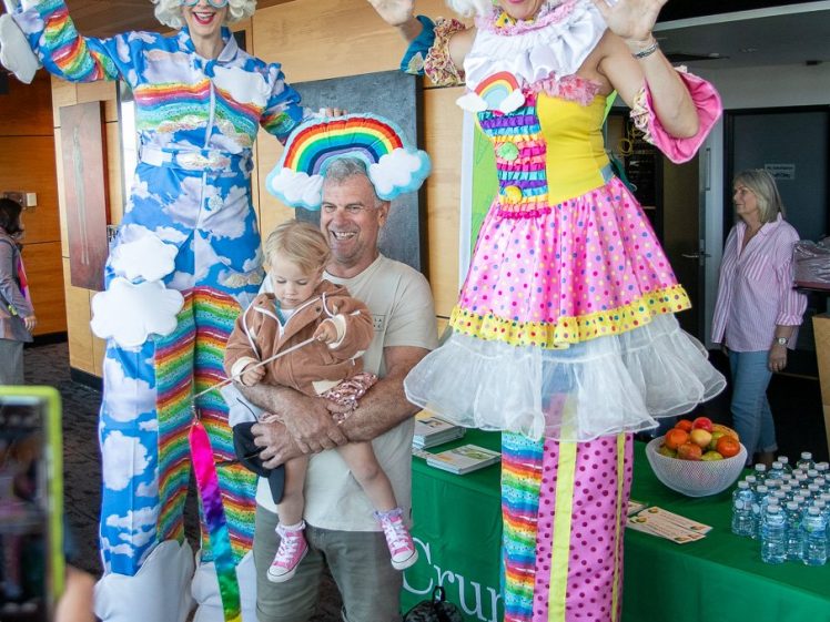 Image of A man holding a young child poses smiling with two colourful performers on stilts dressed in rainbow and cloud-themed costumes at an indoor event. A table with fruit and water bottles is in the background.