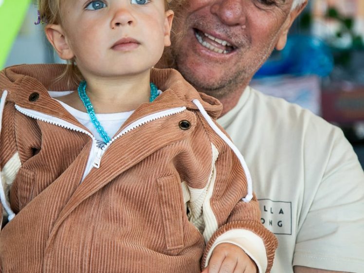 Image of An older man smiles whilst holding a young girl with blonde hair, dressed in a brown jacket and a pink sequin skirt. The girl looks away thoughtfully, whilst the man is looking at her fondly.
