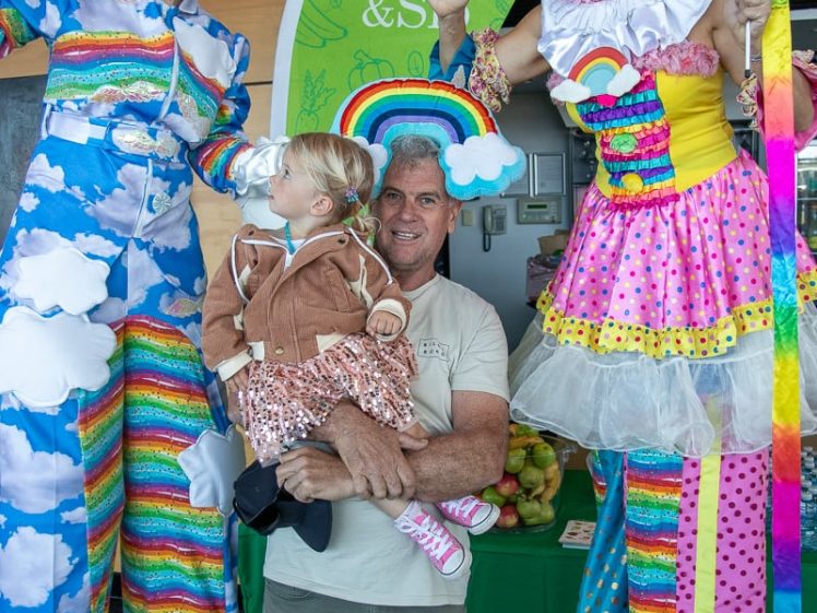 Image of A man holding a young girl poses with two smiling performers on stilts, dressed in colourful, cloud-themed costumes with rainbow accessories, in front of a green Crunch & Sip sign at an indoor event.
