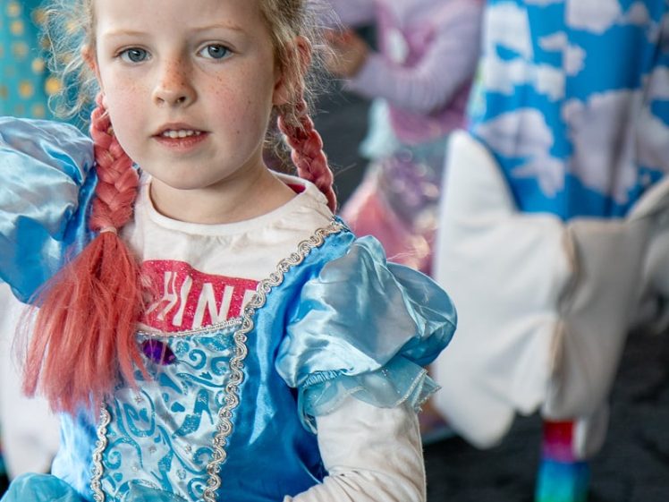 Image of A young girl in a blue princess costume with pink plaits stands in focus, holding her dress. Behind her, other children in colourful costumes are visible, one dressed as a unicorn with a cloud-patterned outfit.