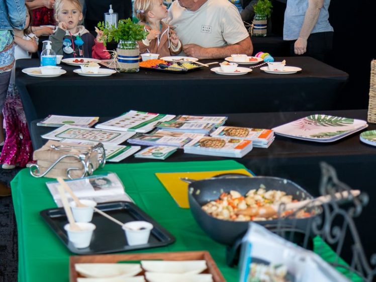 Image of A group of adults and children gather around tables at an indoor event, sampling food and looking at informational leaflets. A woman in a colourful hat stands near a display of dishes and recipe cards in the foreground.