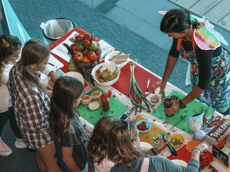 Image of A woman in fairy wings demonstrates food preparation at a colourful table to a group of children, with bowls of chopped vegetables, tomatoes, and ingredients displayed. The setting appears to be a classroom or event space.