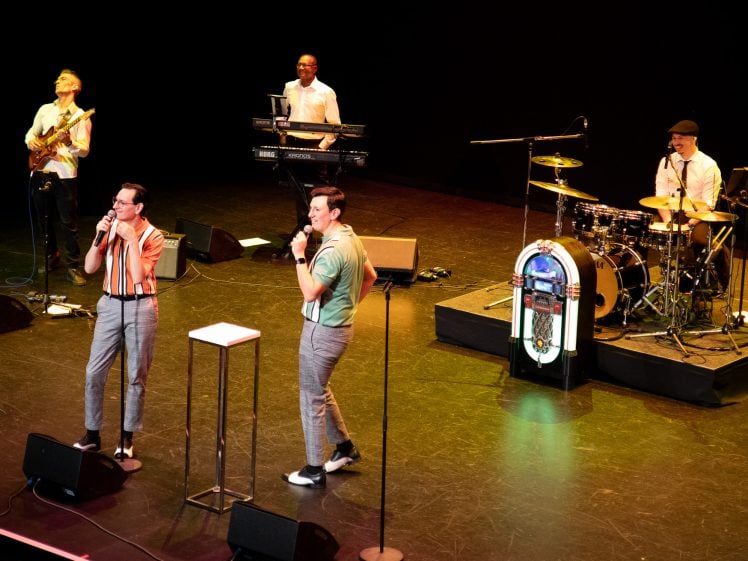 Image of Five musicians perform on stage: two men sing at the front, whilst three musicians play bass, keyboard, and drums. A colourful jukebox stands near the drums. The stage is brightly lit against a dark background.