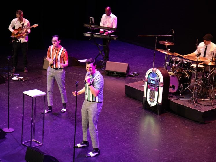 Image of A five-member band performs on stage with two singers at the front, one drummer, one keyboard player, and one guitarist. A jukebox stands near the drum kit, and the musicians wear vintage-style outfits.