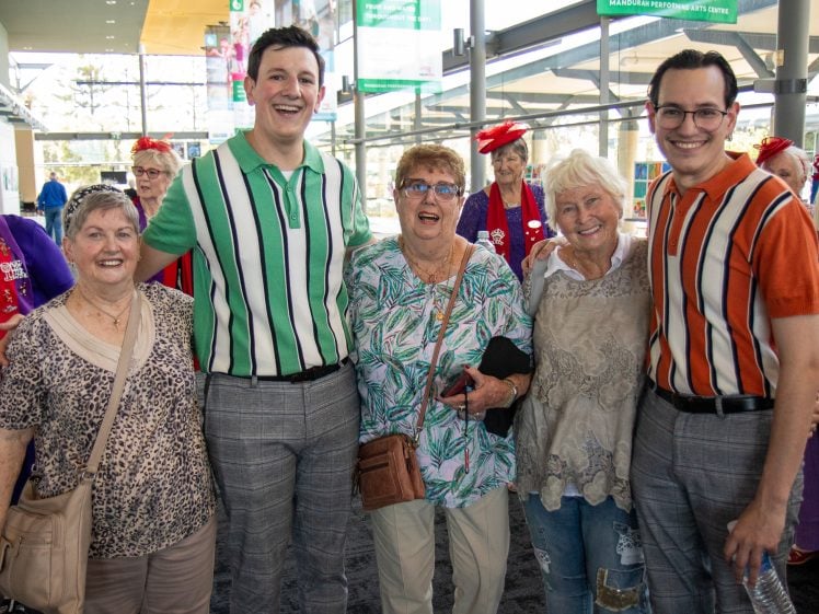 Image of A group of five smiling people, including three older women and two younger men in striped shirts, pose together indoors. Several more people, some wearing red hats, stand in the background.