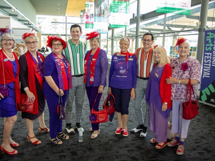 Image of A group of eight smiling adults, mostly older women wearing red and purple outfits with hats, and two men in striped shirts, pose together indoors at the Mandurah Arts Festival.
