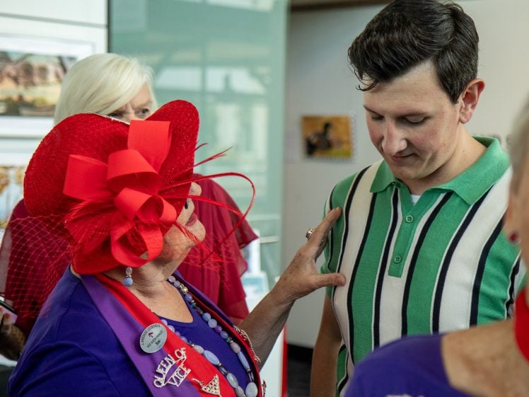 Image of An older woman in a bright red hat and purple outfit speaks to a young man in a green striped shirt whilst touching his arm in a social setting with other people in the background.