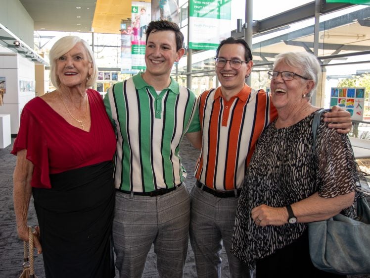 Image of Four people stand together indoors, smiling at the camera. Two older women are on either side of two younger men wearing striped shirts. Artwork and large windows are visible in the background.