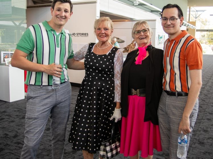 Image of Four people stand indoors, smiling at the camera. Two women wear vintage-style dresses—one in black polka dots and one in bright pink. Two men wear striped shirts and hold water bottles. Art and large windows are visible in the background.