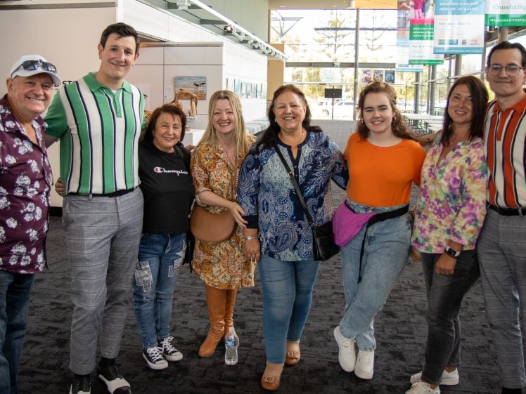 Image of A group of eight people stand close together indoors, smiling at the camera. They wear colourful, casual clothes, and the background shows windows, a carpeted floor, and some informational signs.
