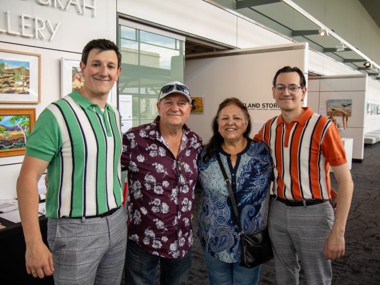 Image of Four people stand smiling together in an art gallery, with two young men in striped shirts standing on either side of an older man in a floral shirt and an older woman in a blue patterned blouse. Framed artwork hangs on the walls behind them.