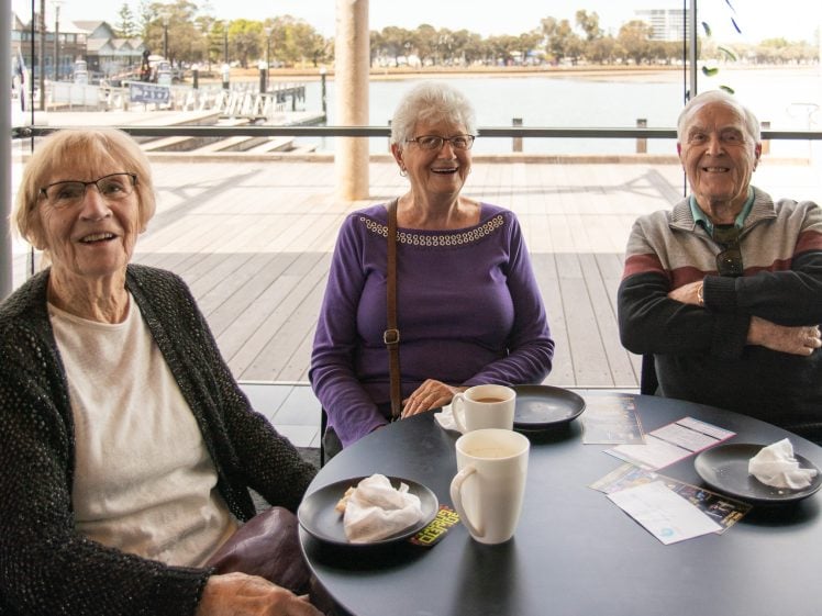 Image of Three elderly people sit at a round table in a café, smiling at the camera. There are coffee cups, plates with crumbs, and leaflets on the table. Large windows behind them reveal a waterfront and trees.
