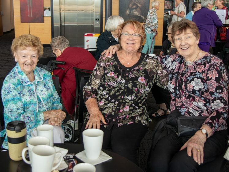 Image of Three elderly women sit together at a table, smiling for the camera. They are indoors, surrounded by other people. Several cups and a flask are on the table in front of them.