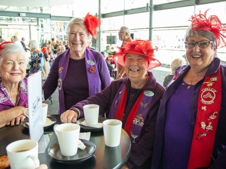 Image of Four older women smile at a table in a bright café, each wearing purple outfits with red hats or accessories. They have cups of coffee and appear to be enjoying a cheerful social gathering.