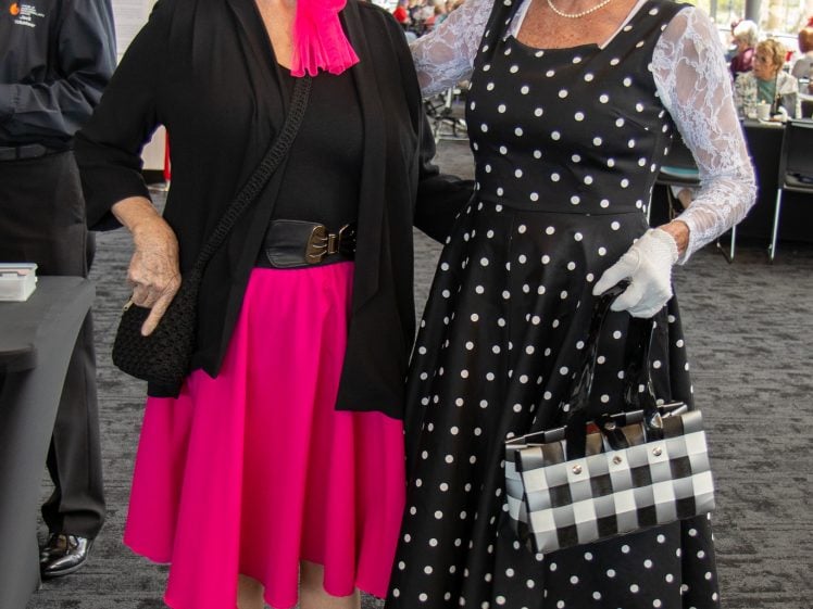Image of Two older women pose together, smiling. One wears a bright pink skirt, black top, and sunglasses; the other wears a black and white polka dot dress, white gloves, and carries a chequered handbag. Both look cheerful and stylish.