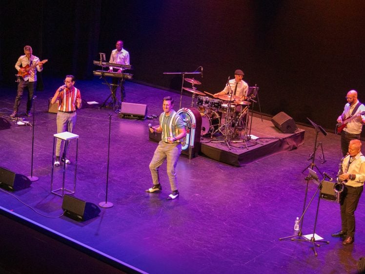 Image of A six-member band performs on a stage under colourful lighting, with two vocalists at the front and four musicians playing guitar, keyboard, drums, and bass in the background.
