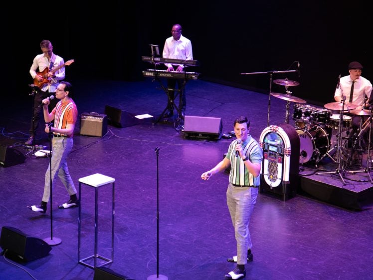 Image of A band performs on stage with two singers in colourful shirts at the front, a guitarist and keyboardist in the back, and a drummer playing a drum kit decorated with a jukebox image.
