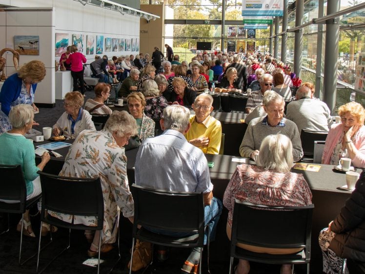 Image of A large group of older adults sit at tables in a bright, modern café or gallery, chatting and having coffee. Artworks and sculptures are displayed on the walls and around the room. Sunlight streams through large windows.