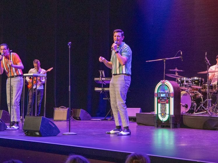 Image of Four musicians perform on stage: two singers at microphones, a guitarist, and a drummer beside a vintage jukebox prop. The performers wear retro clothing, and the stage is lit with colourful lights.