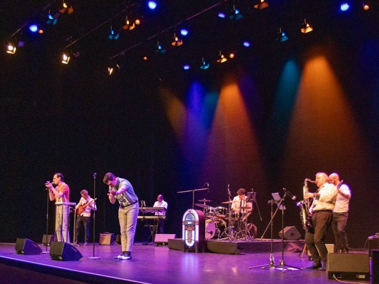 Image of A band performs on stage under colourful lights, featuring musicians playing guitar, keyboard, drums, saxophone, and trumpet, with two singers at the front. A jukebox prop is near the drummer. The background is dark.
