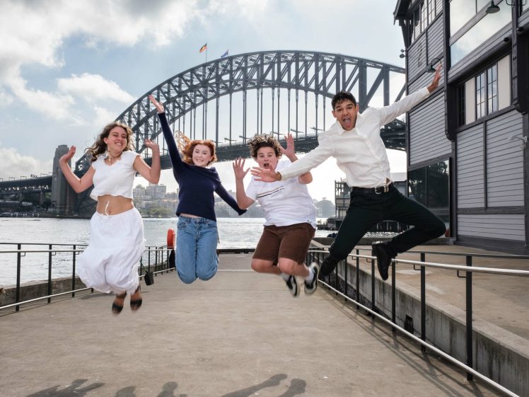 Image of Four young people joyfully jump in the air with the Sydney Harbour Bridge in the background on a cloudy day, near the waterfront and beside a modern building.