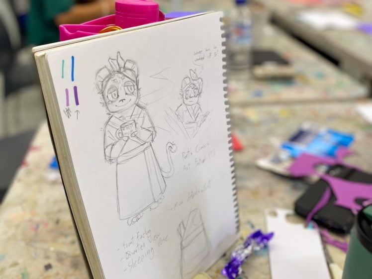 Image of A sketchbook on a cluttered table shows drawings of a cartoon cat character in different poses and expressions. Art supplies and paper surround the sketchbook, and people are working in the blurred background.