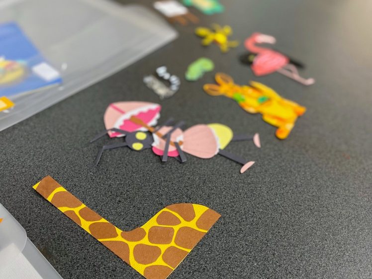 Image of Colourful paper cut-outs of various animals, including a giraffe, ant, rabbit, flamingo, and frog, are arranged in a line on a grey tabletop. The focus is on the giraffe-patterned piece in the foreground.