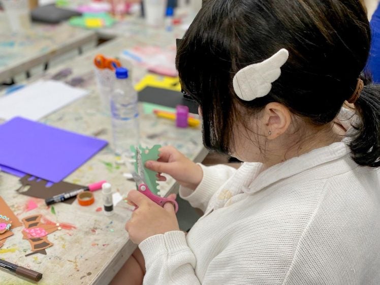 Image of A child with dark hair and a white hair clip uses craft scissors to cut green paper at a table covered with art supplies and colourful paper. Other children are blurred in the background, working on crafts.