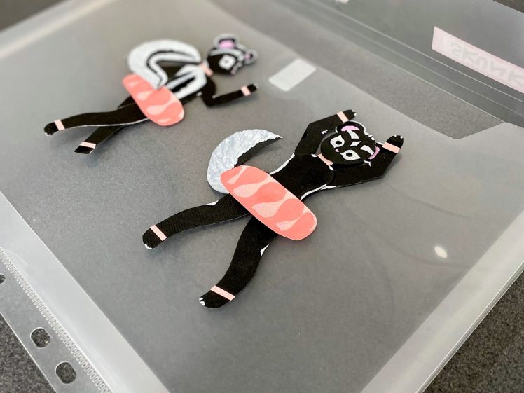 Image of Two handmade paper cut-outs of skunks with sushi rolls on their bodies are placed inside a transparent plastic wallet on a grey surface. The skunks are black with white stripes and have pink sushi accessories.