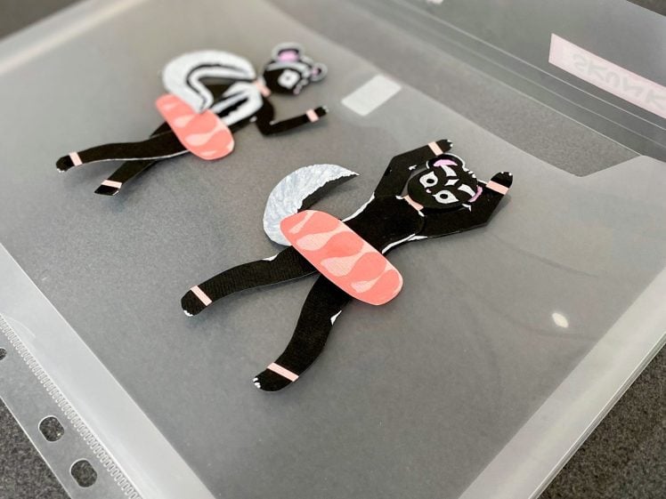 Image of Two flat, cartoon-style skunk characters made from cut paper, each with a sushi roll as their midsection, are displayed on a translucent plastic folder.