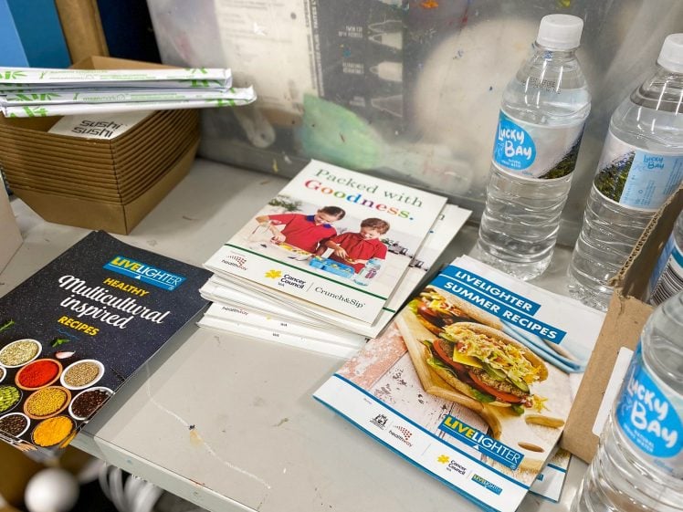 Image of A stack of recipe booklets, including Healthy Multicultural Inspired Recipes and LiveLighter Summer Recipes, sits on a table next to sushi boxes and several bottles of water labelled Lucky Bay.