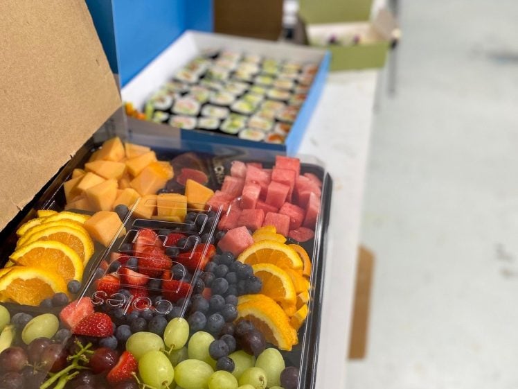 Image of A tray of assorted fresh fruit, including grapes, oranges, strawberries, blueberries, and melon, sits on a white table next to a box of sushi rolls and bottled water in the background.
