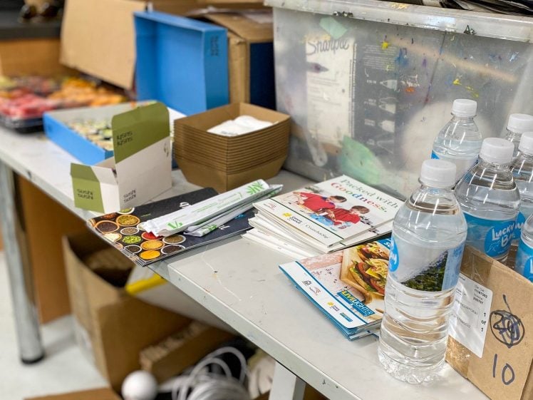Image of A table with bottled water, food brochures, wooden cutlery, serviettes, and boxes, with various containers and supplies in the background. A box labelled Lucky Bay with the number 10 is also visible.