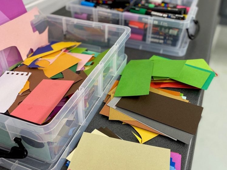 Image of Containers filled with colourful paper scraps and art supplies are arranged on a long table, ready for a crafting activity. Various shades of paper are spread out on the table in an organised yet creative manner.