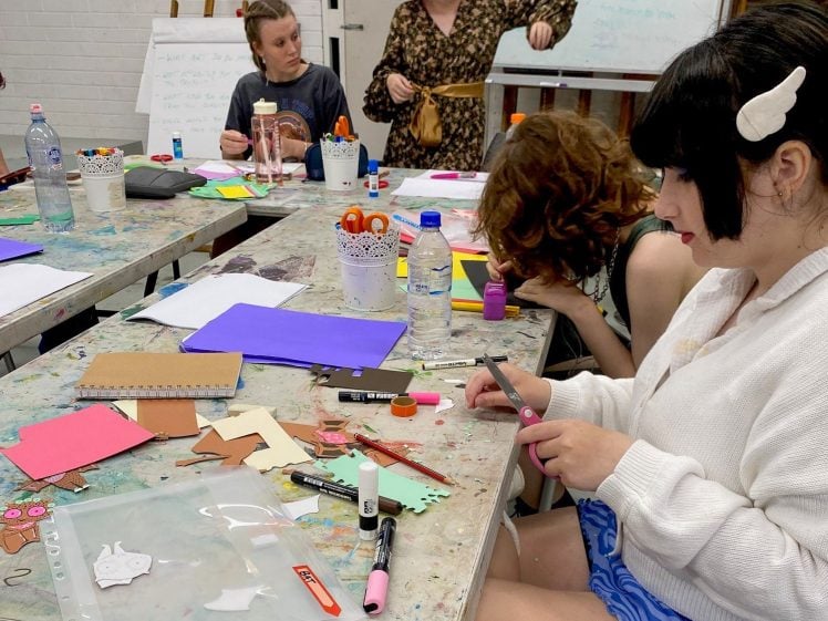 Image of Four people sit at messy tables covered with craft supplies, paper, and felt-tip pens, working on art projects. One person stands in a peach hat, while others are focused on cutting or assembling materials. Whiteboard in background.