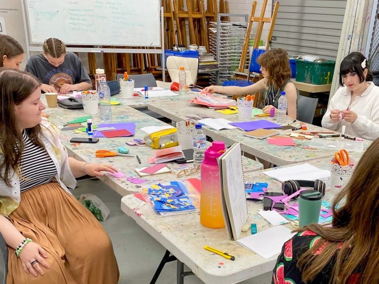 Image of A group of young people sit around a large table covered with colourful craft materials, paper, and art supplies, working on creative projects in a bright classroom or art studio.